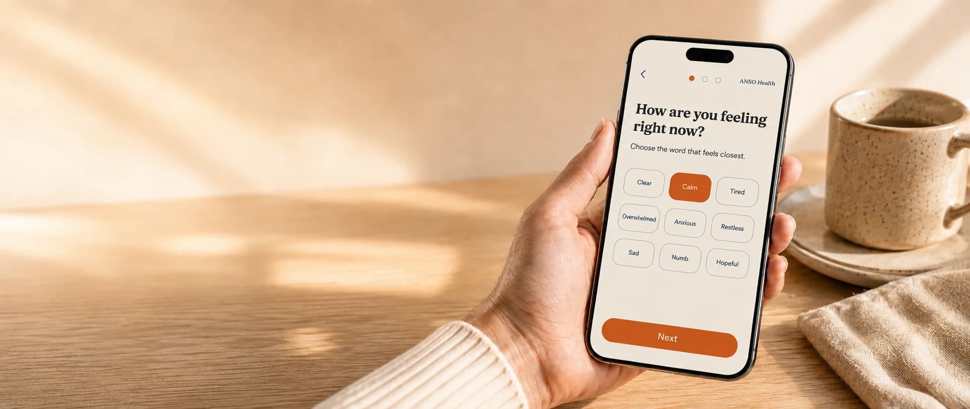 Mano sosteniendo un teléfono con el check-in de ANSO Health: la pregunta de cómo te sientes ahora y una grilla de palabras para elegir, junto a un café sobre un escritorio soleado